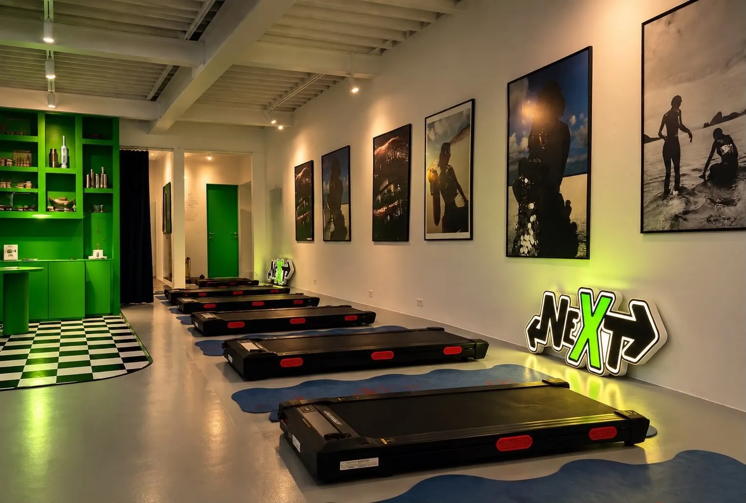 Salle de treadmill du club NEXT : une rangée de tapis de marche noirs, des photos d’art encadrées au mur et l’enseigne lumineuse NEXT au sol